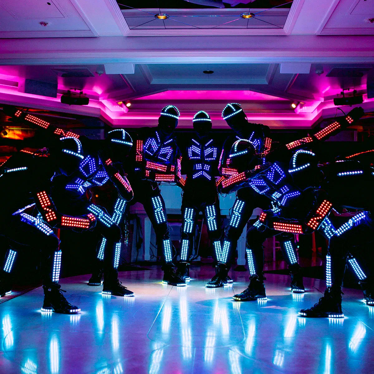 Tron dancers in black suits with glowing blue and red LED strips and LED helmets striking a pose on a shiny dance floor under pink ceiling lights