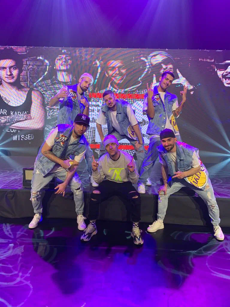 Six young men posing on a stage in front of a big LED screen under purple stage lights – the Tron dancers in matching denim vests, some in caps, with the choreographer in a grey sweatshirt and beanie crouching in the middle