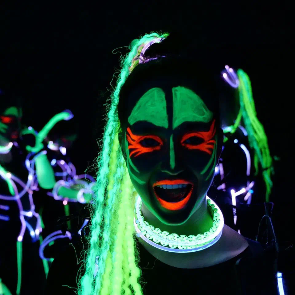 A Tron Girl with neon green UV face paint, orange eye make-up, glowing green braids and a glowing necklace, laughing in the dark, with other dancers in glowing outline costumes behind her