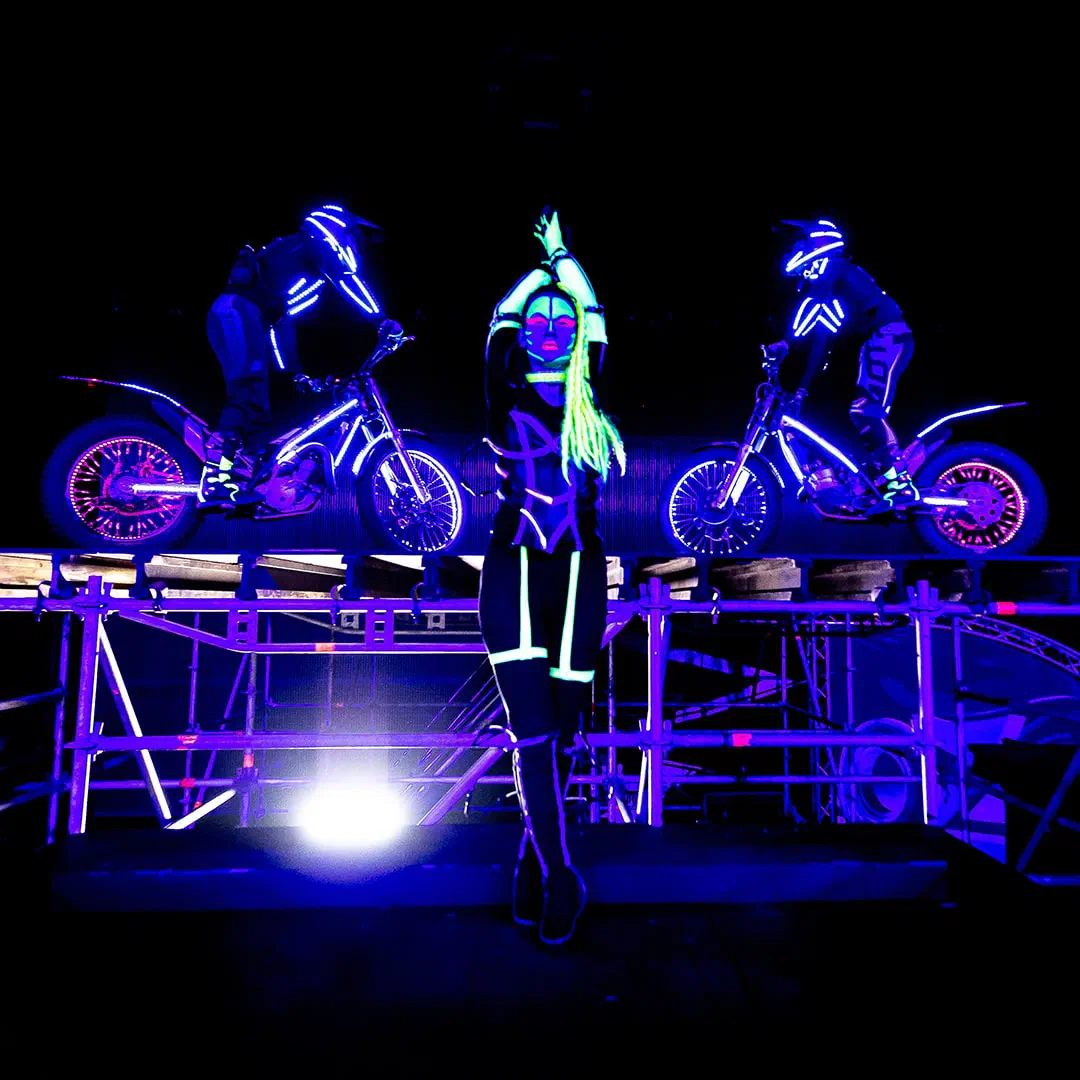 Tanečnica Tron Girls s UV líčením a neónovo zelenými vrkočmi v čiernom kostýme so žiariacimi líniami pred dvoma motorkármi v prilbách a kombinézach s LED osvetlením na pódiu z lešenia