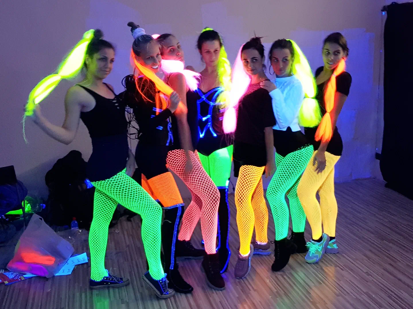 Seven dancers under UV light in a rehearsal room, in glowing neon green, pink, orange and yellow fishnet tights with neon braids, two of them with blue fiber optic lines on their black tops