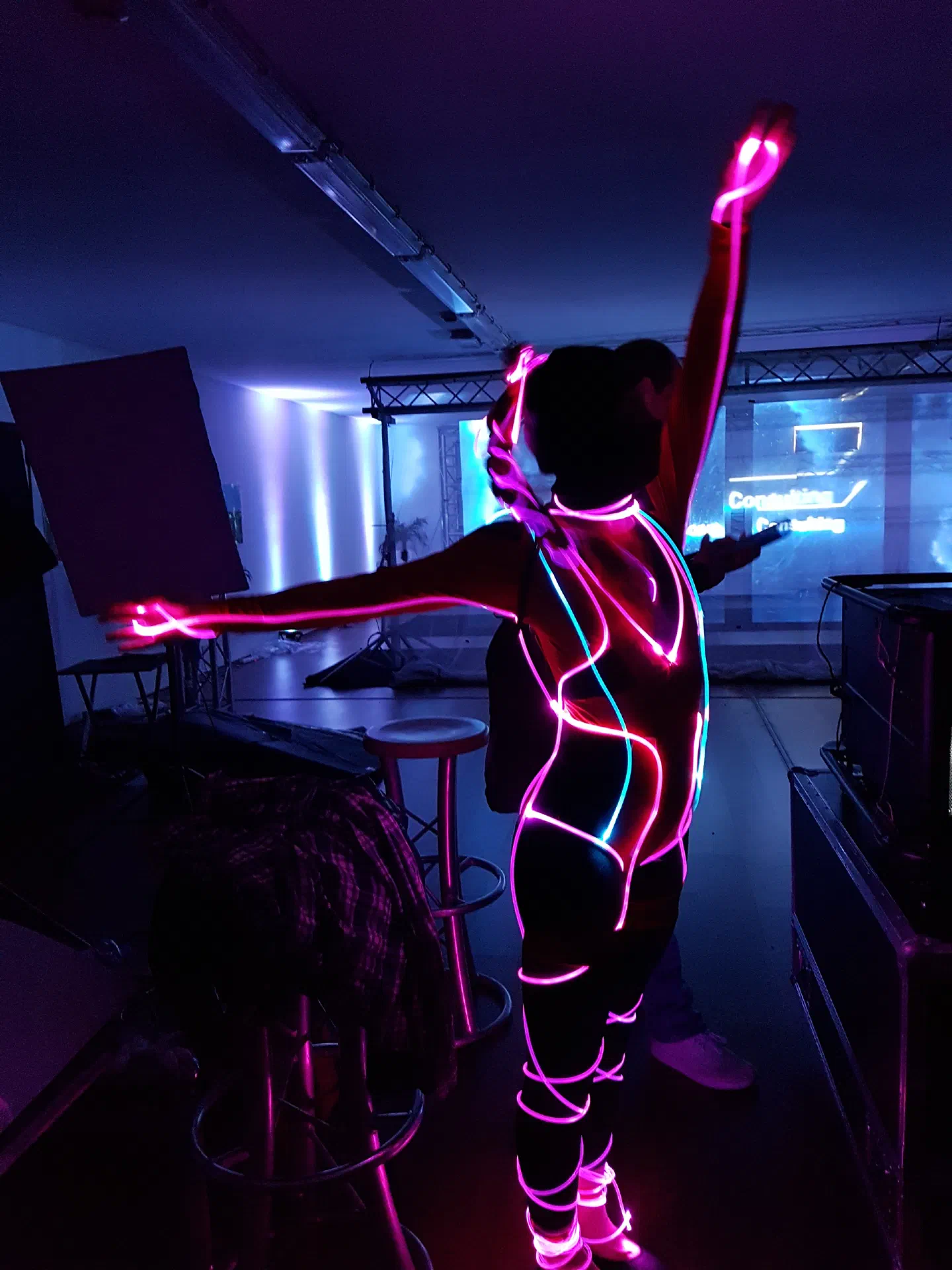 A dancer with one arm raised and the other stretched out, in an early Tron Girls costume outlined with glowing pink and blue fiber optic cable, in a dim hall with a lighting truss and a projection screen