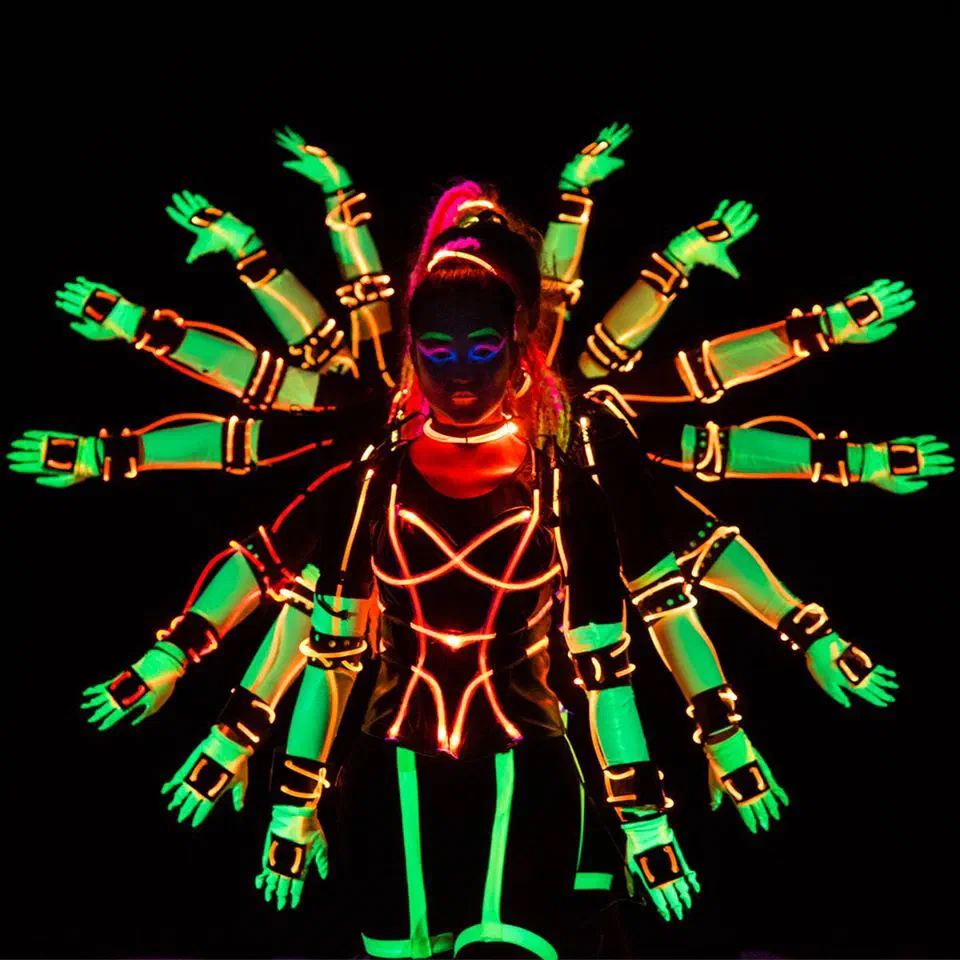 A Tron Girl with neon UV make-up and pink braids in a suit outlined with orange fiber optic cable, the neon green arms of the dancers behind her fanned out around her