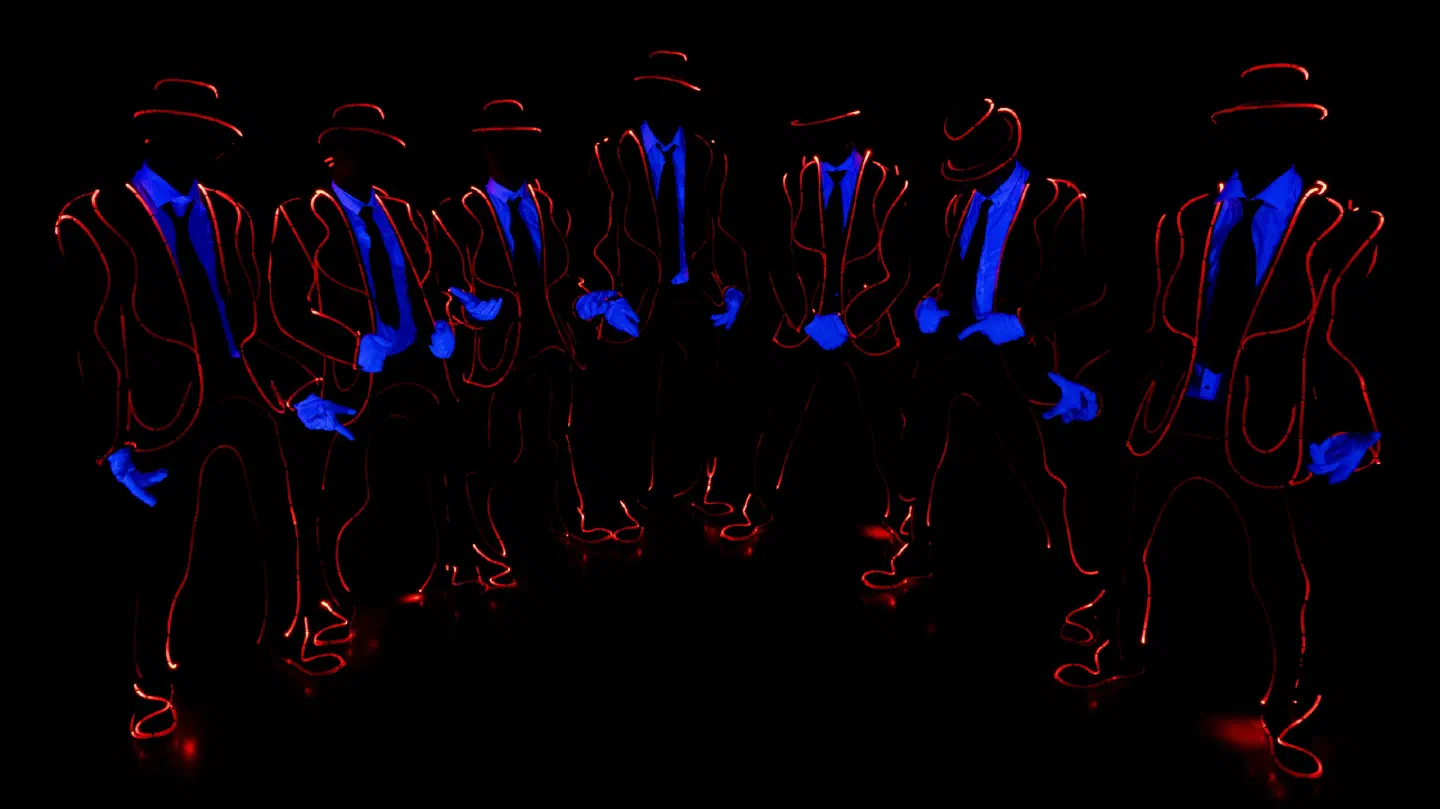 Seven Tron Man dancers in hats and suits outlined with red fiber optic cable, their gloves and tie silhouettes glowing blue under UV light