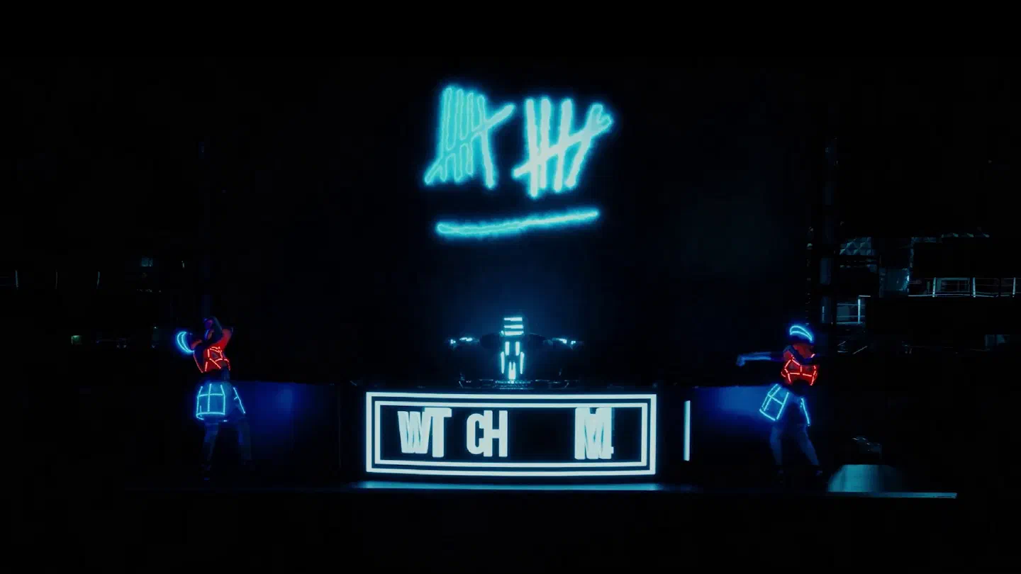 Tron DJ in a glowing LED helmet at a DJ booth with a white LED frame, a blue laser logo above him and two dancers in red and blue LED suits on both sides of the dark stage