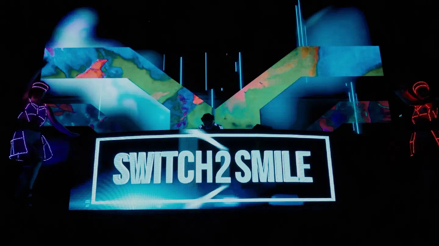 DJ Switch2Smile behind a booth whose LED screen shows the Switch2Smile logo, colourful LED wall visuals around him and dancers in LED suits on both sides