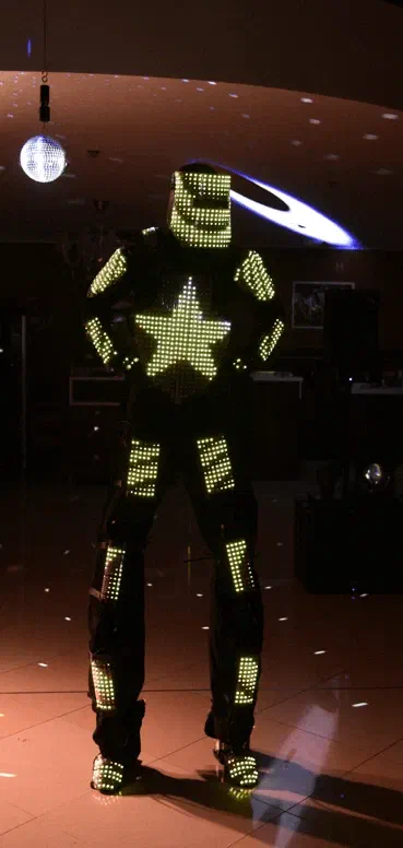 Tron Pixel Robot stojí na tanečnom parkete pod diskoguľou, jeho LED kostým žiari žltozeleno s veľkou hviezdou na hrudi