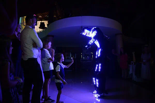 Deti a svadobní hostia sledujú Tron Pixel Robot žiariaci bielymi a modrými pixelmi v tmavej sále, jeden chlapec sa k nemu načahuje