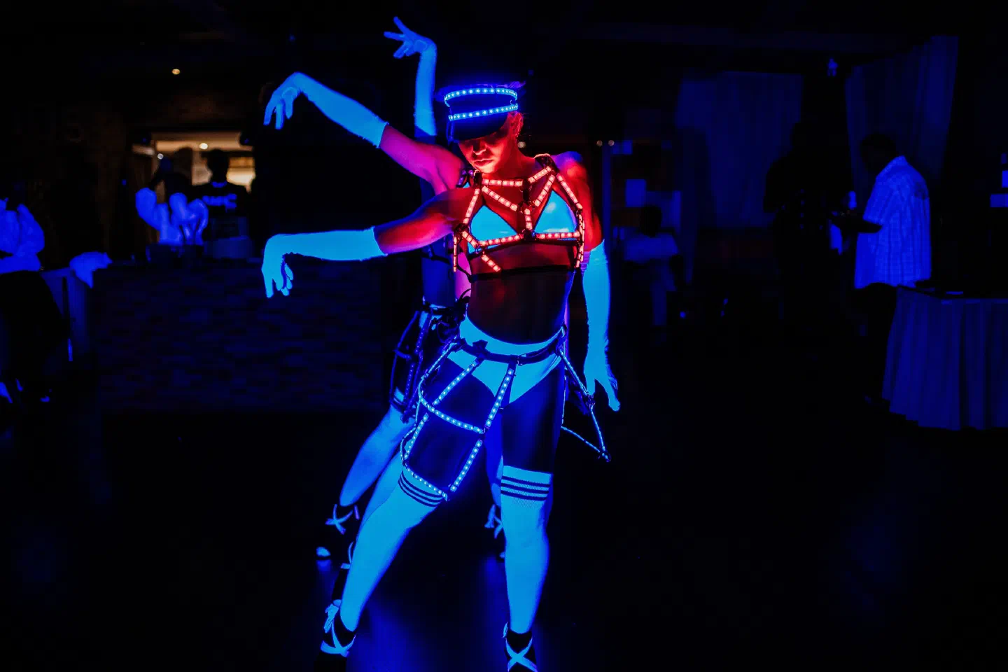 Dve tanečnice Tron Dolls pod modrým svetlom v tmavej svadobnej sále, predná v čiapke a postroji lemovanom červenými a bielymi LED, tá za ňou dvíha ruky, v pozadí hostia