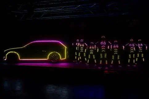 The outline of a Volvo XC90 lit by pink and yellow LED strips next to a line of Tron dancers in glowing LED suits on a dark stage