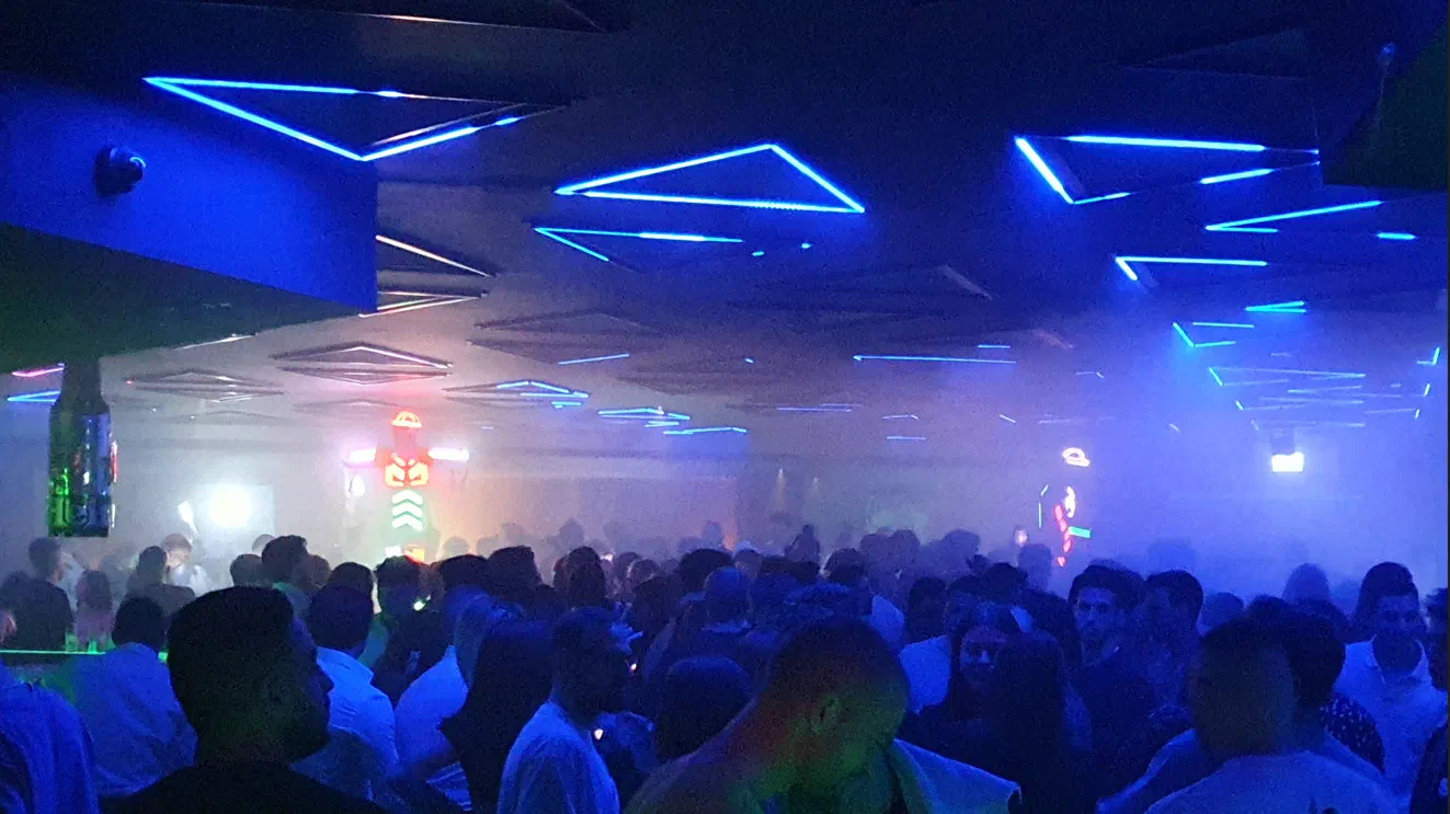 Široký záber na preplnený nočný klub s modrými svetelnými LED trojuholníkmi na strope a dvoma tanečníkmi v žiariacich červených a zelených LED kostýmoch, ktorí sa týčia nad davom v opare