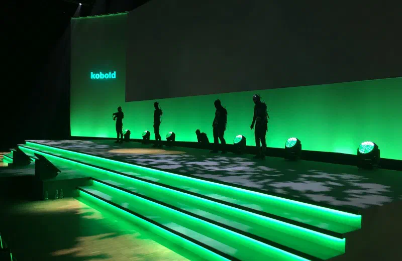 Tron dancers in unlit LED suits on a stage lit in green, with the kobold logo on the wall behind them and glowing LED steps in front