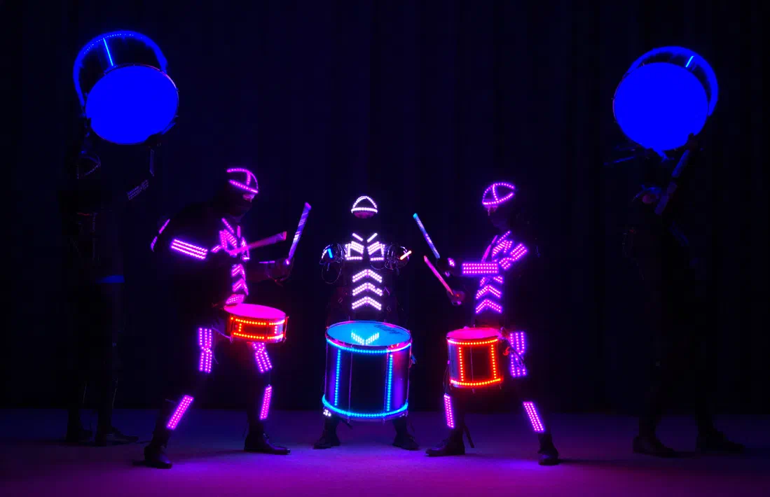 Five LED drummers on a dark stage – two in pink LED suits and one in a white LED suit playing LED-lit drums in the middle, two holding big blue glowing drums up high on the sides