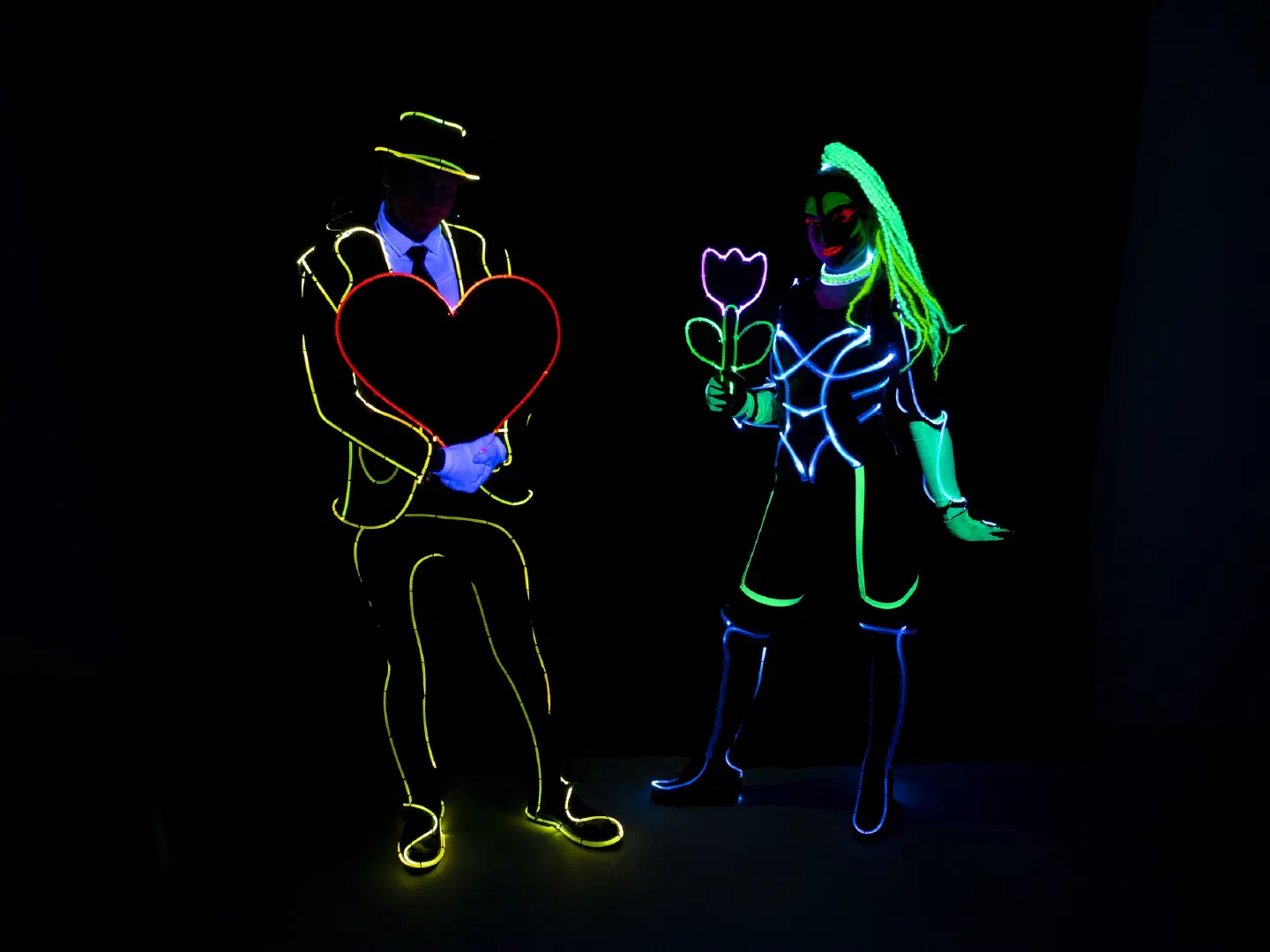 A dancer in a glowing yellow outline suit and hat holds a red LED heart next to a dancer in a blue and green glowing UV costume holding a glowing flower