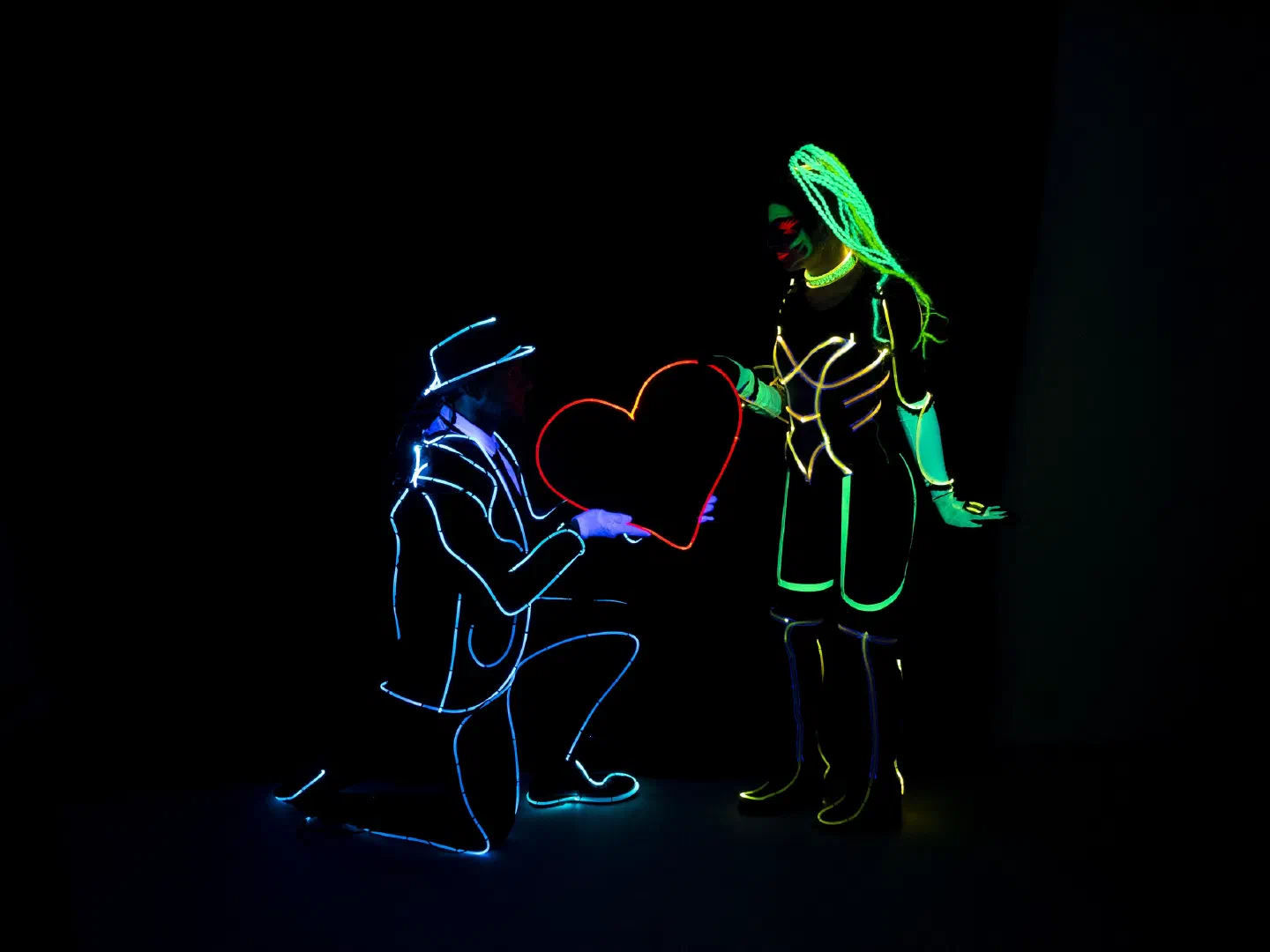 A dancer in a glowing blue outline suit and hat kneels and offers a red LED heart to a dancer in a green and yellow glowing UV costume with neon braids
