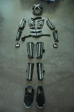 Parts of the LED Suit v.2 laid out on a concrete floor – black plastic shells for the head, chest, arms and legs lined with LED strips, and a pair of shoes
