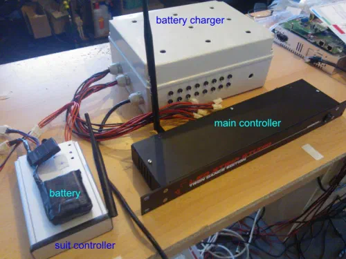 Version 2 LED dance controllers on a workbench – a rack-mount main controller with an antenna, a suit controller with its battery and a battery charger box