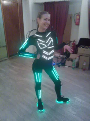 Smiling dancer in a black suit with glowing white and turquoise LED strips on the chest, arms, legs and shoes, posing in a rehearsal room