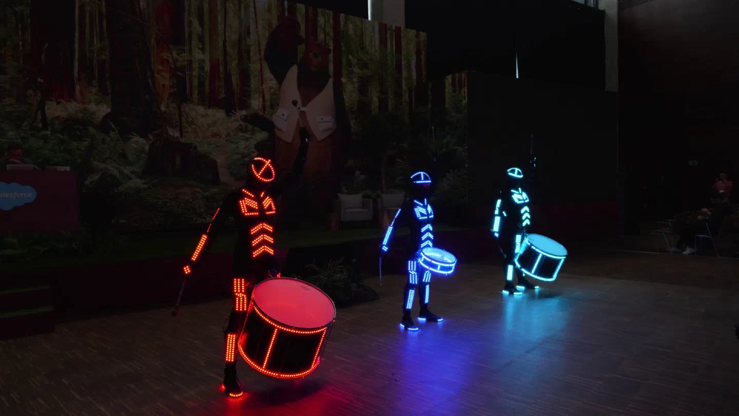Three Tron Drummers in red, blue and turquoise LED suits with matching LED drums on a dark stage in front of a forest backdrop