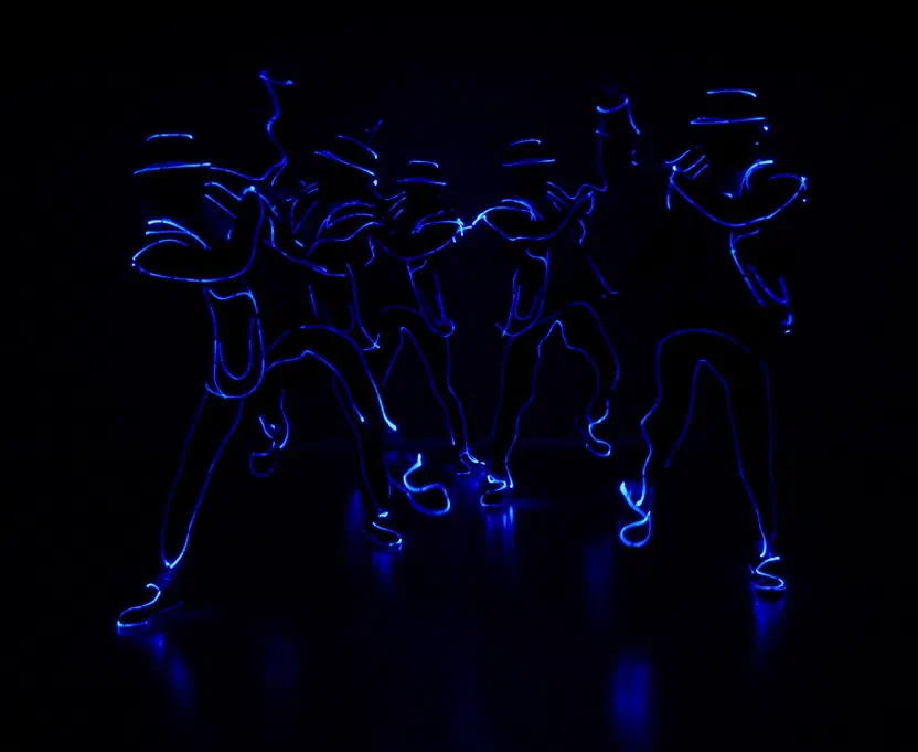 Five Tron Man dancers in hats and suits outlined with glowing blue lines, leaning forward together in a dark room