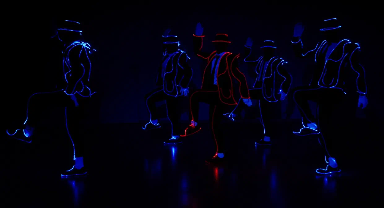 Five Tron Man dancers in hats and suits outlined with glowing blue lines – the one in the middle in red – in a Michael Jackson knee-up pose in the dark