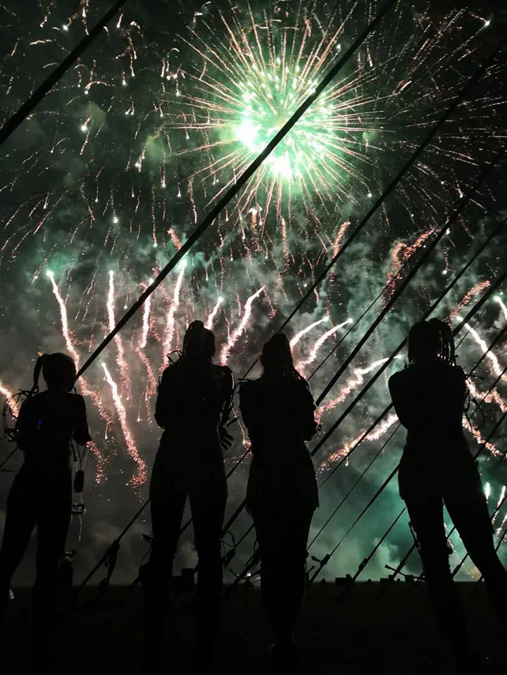 Silhouettes of four Tron Girls with braided hair in their wired LED costumes, watching a huge green and gold fireworks display in the night sky behind diagonal rigging cables