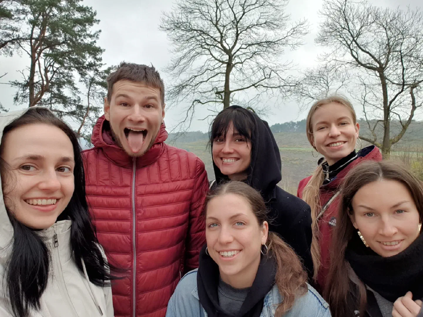 Selfie of our UK team – five smiling young women and a young man in a red puffer jacket sticking out his tongue, with bare trees and green hills behind them on a cloudy day