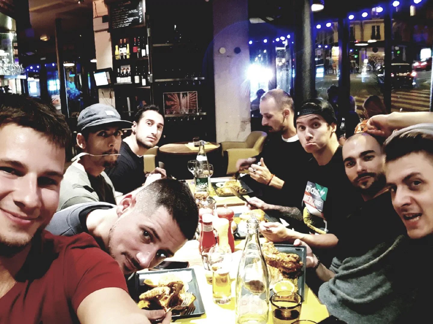 Selfie of our team – eight young men at a long table in a Paris restaurant at night, with plates of food, bottles and glasses, and a lit street behind the windows