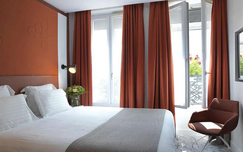 Room in the Hotel Vendôme – a double bed with white sheets and a grey throw, rust-coloured curtains and an armchair by the open balcony window