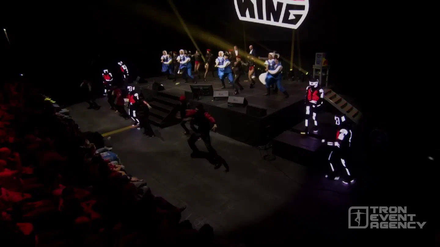 Finale of the EisKing opening ceremony – folklore dancers in blue costumes and dancers in red on the stage, Tron dancers in glowing LED suits around it, the audience in the foreground