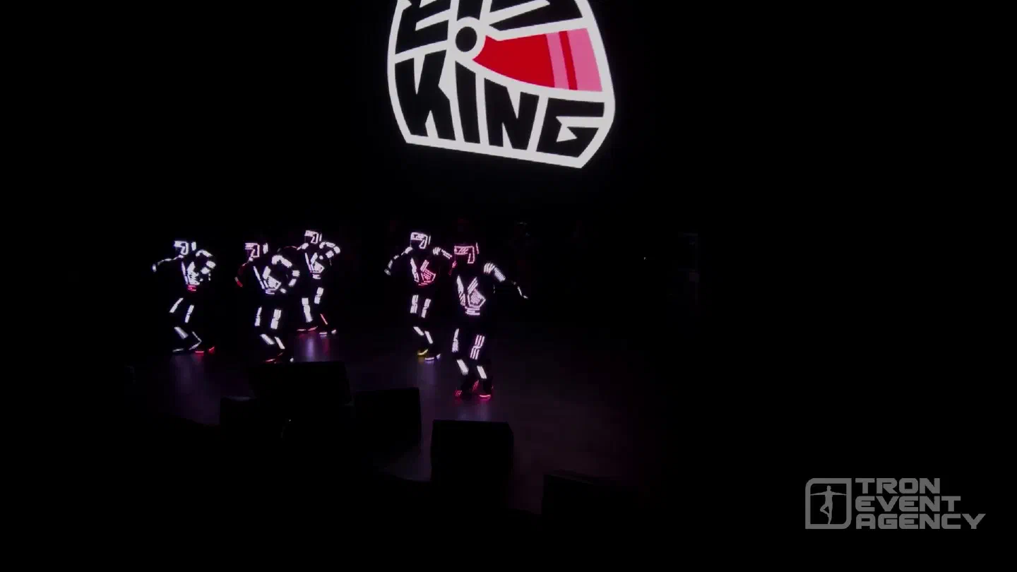 Five Tron dancers in white and pink LED pixel suits dancing on a dark stage under the glowing EisKing helmet logo