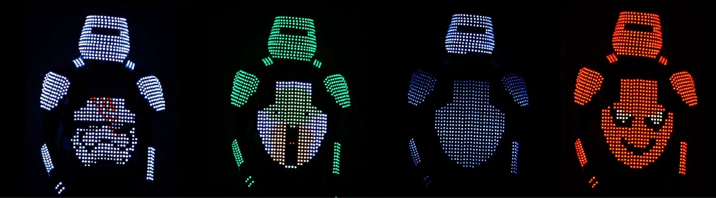 Four Pixel Tron Dance suits in the dark, seen from the front – the pixels on the helmets, shoulders, arms and chests glow white, green, blue and red, the red chest showing a smiling face