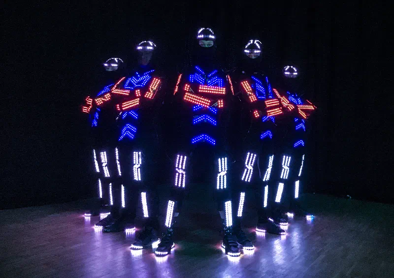 Five dancers in black LED suits and LED-lit helmets standing in a V formation on a wooden floor, with red, blue and white LED strips glowing on their chests, arms and legs