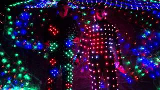 Two dancers in black pixel costumes covered with glowing red, green, blue and white LED dots, in front of a wall of colourful LED pixels
