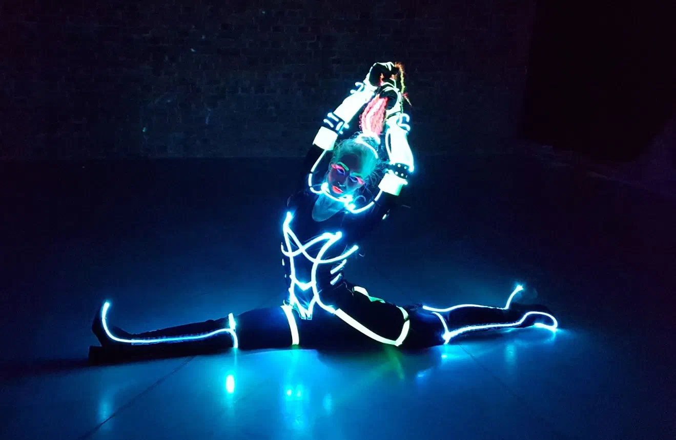 Female dancer doing the splits on a dark floor in a black costume outlined with glowing white-blue light lines, her face paint and braided hair glowing in UV light