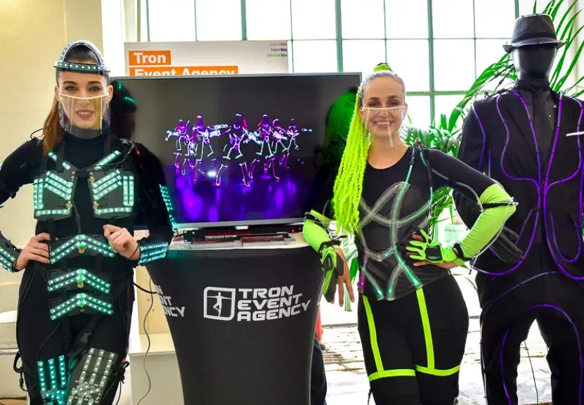Two smiling dancers with clear face shields at our Tron Event Agency stand – one in an LED suit glowing turquoise, one with neon yellow braids and green lines – beside a screen showing LED dancers and a mannequin in a hat with purple lines