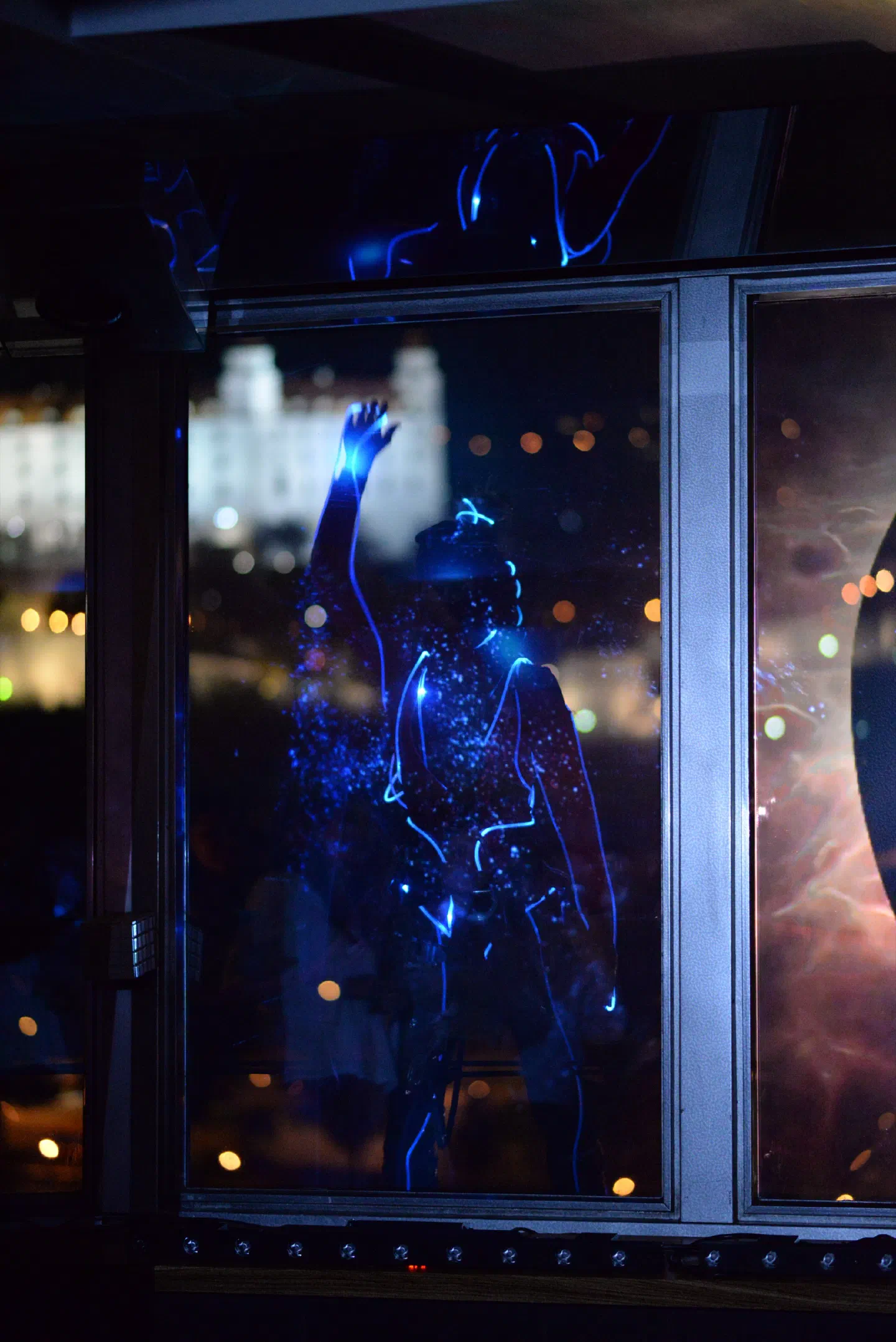 A Tron Girl outlined in glowing blue lines dances with one arm raised behind a window of the UFO restaurant at night, with the lit-up Bratislava Castle in the background