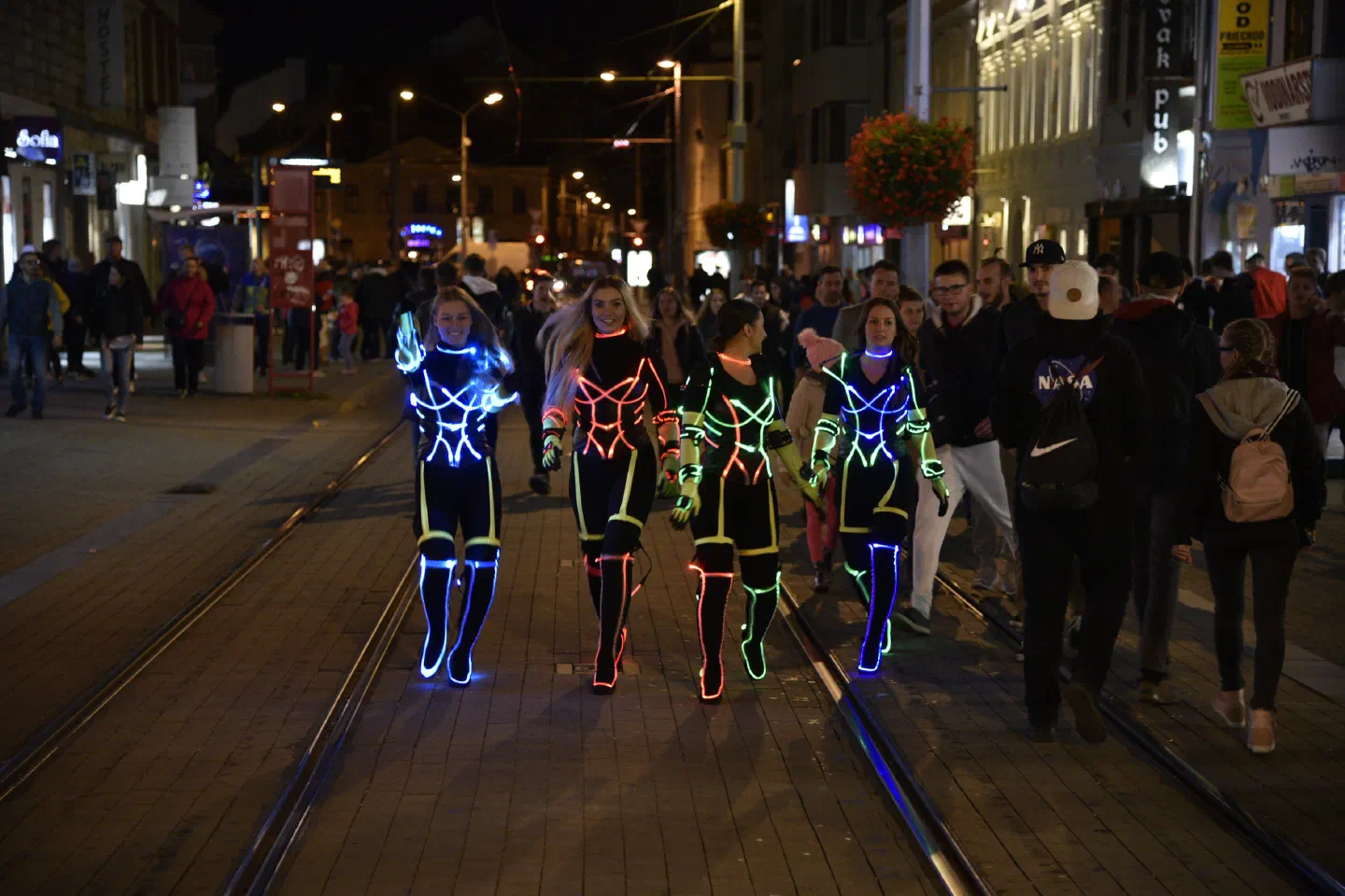 Four Tron Girls in black suits with glowing blue, red and green LED lines walk along the tram tracks of a busy Bratislava street at night, among passers-by