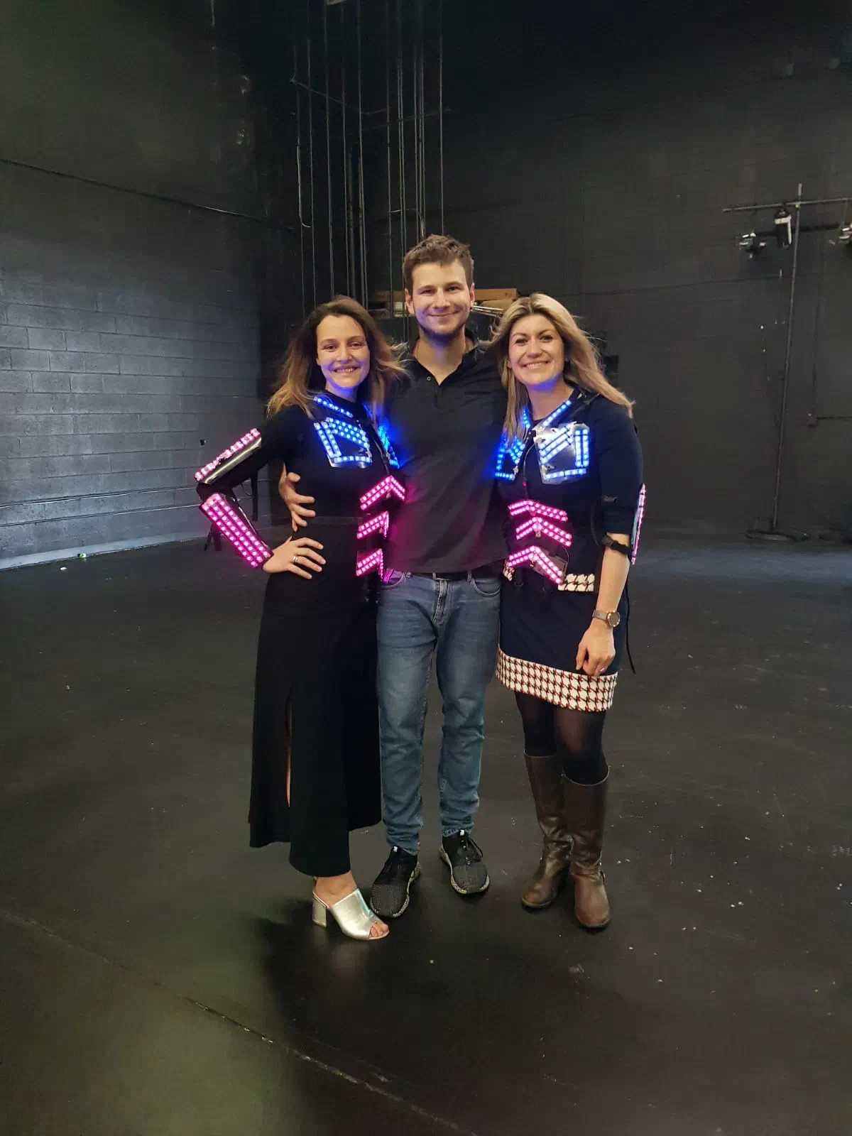 Richie in a dark polo shirt and jeans between two smiling clients who wear the upper parts of our LED suits glowing blue and pink, on a black studio stage