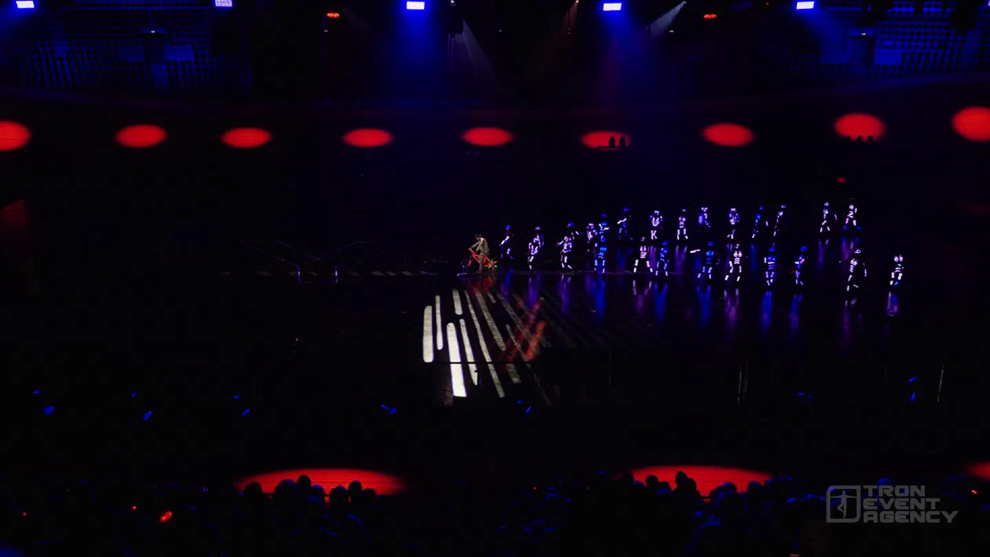 A rider on a bicycle lit with red and green LEDs in front of a long line of dancers in LED suits on the Velodrom floor, white light stripes on the floor and the audience in the foreground