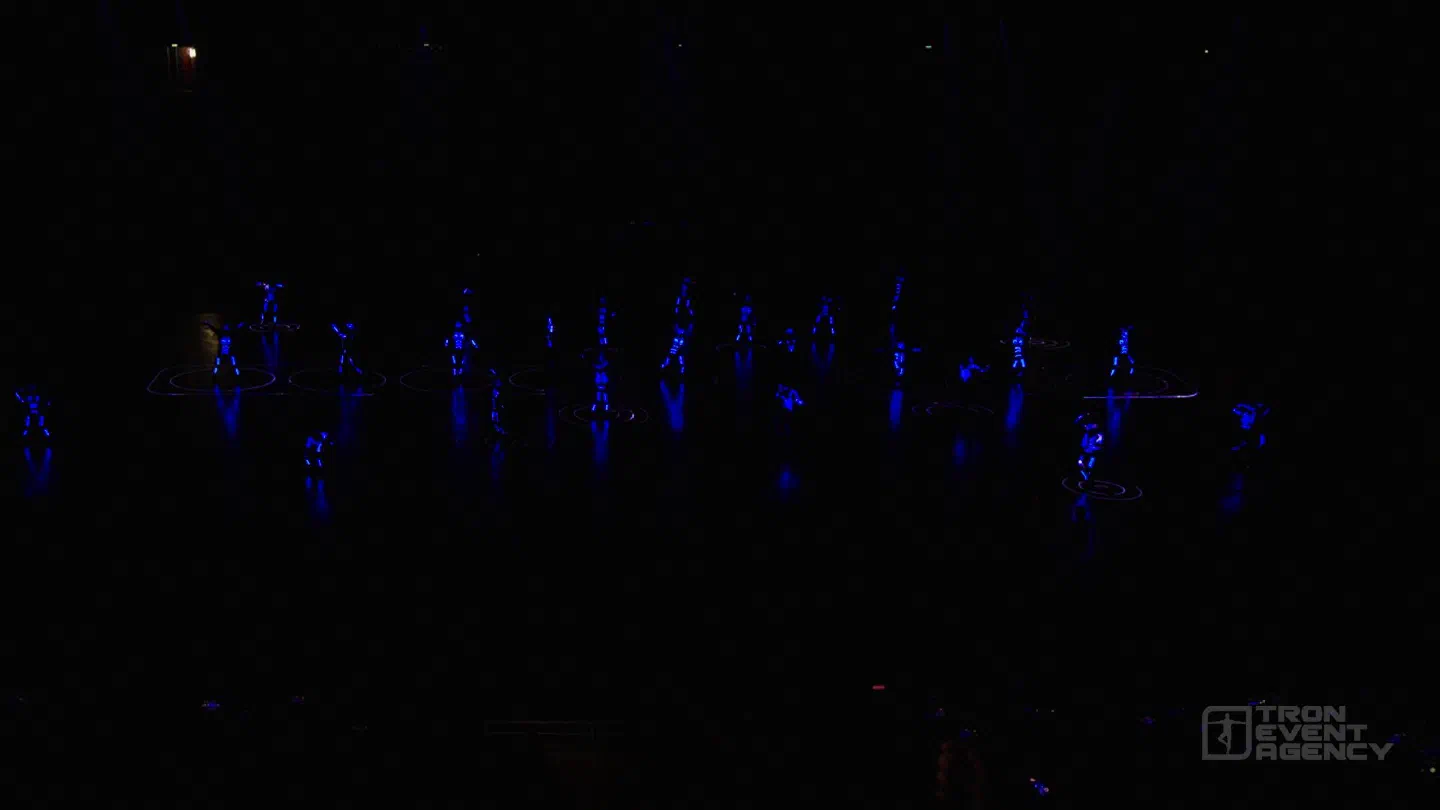 About 25 dancers in blue LED suits spread across a dark stage, standing in circles drawn on the floor by lasers