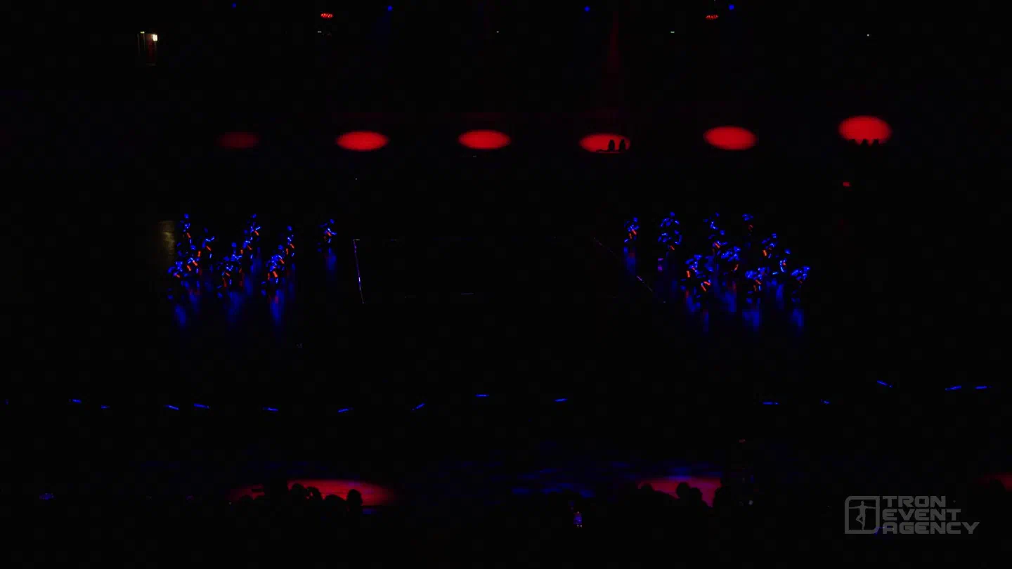 Two groups of dancers in blue and red LED suits on a dark arena floor, red spotlights on the wall behind them and a line of small blue lights in front