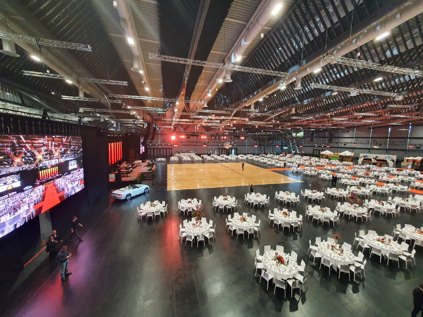 Empty event hall before the show – round banquet tables with white chairs around a wooden dance floor, a white car on display and a large LED screen on the left, lighting trusses under the roof