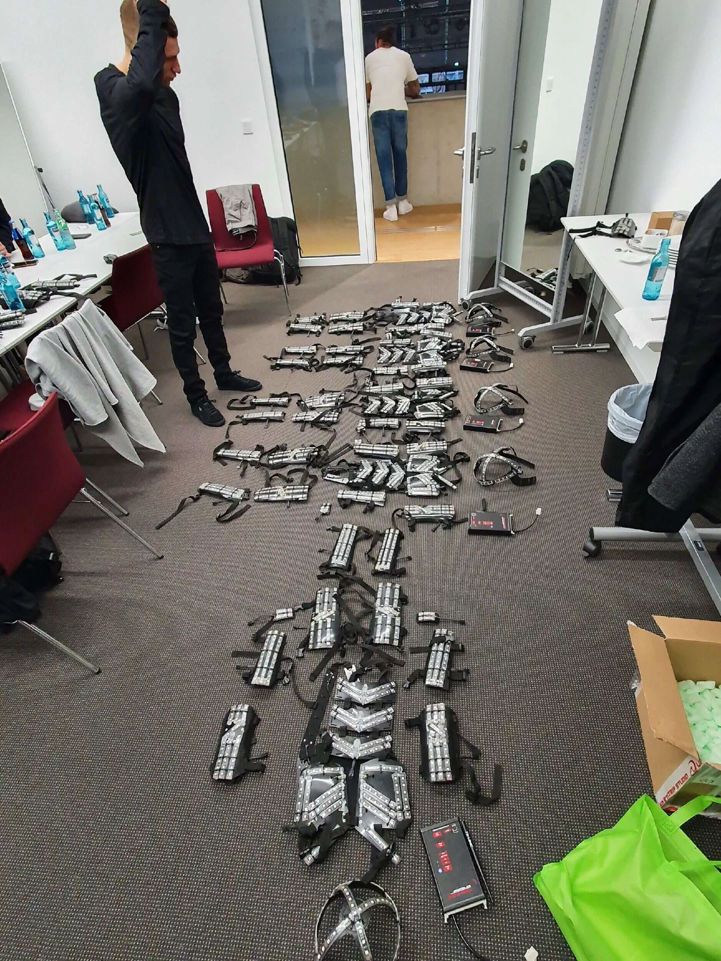 Parts of our Tron Dance LED suits – LED strip panels with straps, LED head harnesses and controllers – laid out in rows on the carpet of a backstage room, a dancer standing beside them