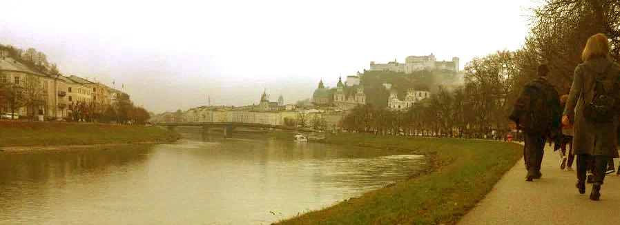 Riverside path along the Salzach in Salzburg on a misty day, with the Hohensalzburg Fortress and the cathedral domes in the distance