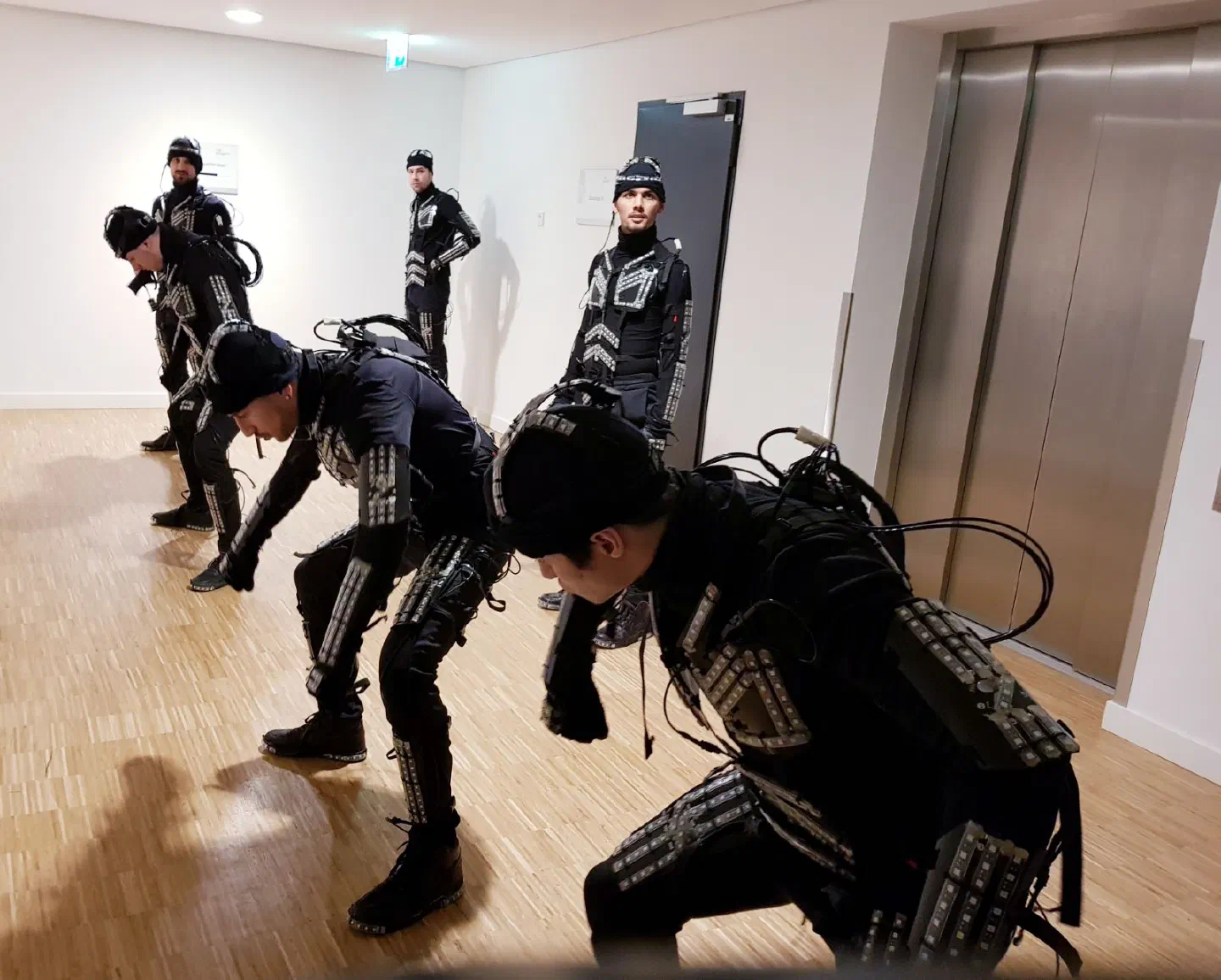 Six Tron dancers in black LED suits, the LED strips switched off, going through the choreography in a corridor with a wooden floor next to a lift