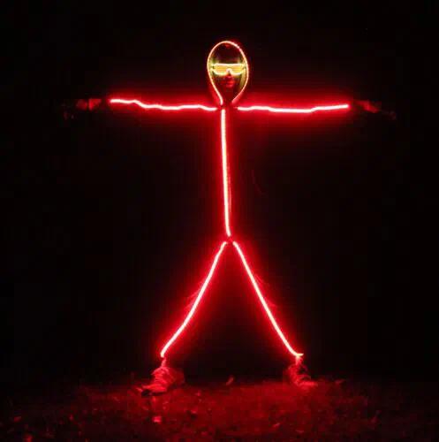 Človek v stickman LED kostýme v noci – červené svietiace línie kopírujú ruky, telo a nohy panáčika a žltá línia obkresľuje hlavu so svietiacimi okuliarmi