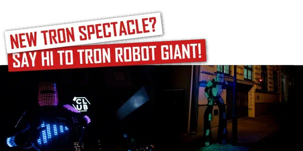 Nová Tron podívaná? Pozdravte Tron Robot Giant! – červené bannery nad Tron Pixel Robotom, LED robotom na chodúľoch, ktorý v noci žiari pixelovými panelmi na mestskej ulici