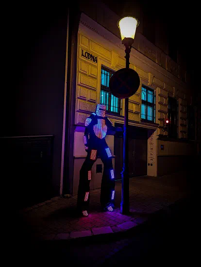 Tron Pixel Robot žiariaci ružovou a modrou sa v noci opiera o pouličnú lampu pred starou budovou