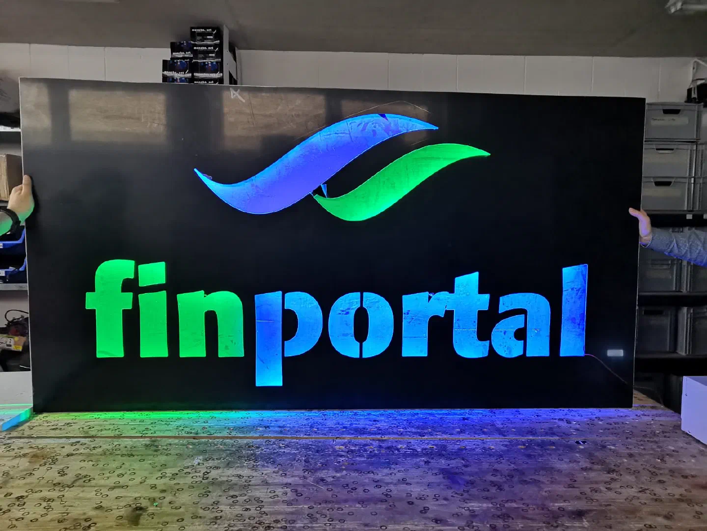 The blue and green Finportal logo glowing as an LED sign on a black board, held upright on a workbench in our workshop